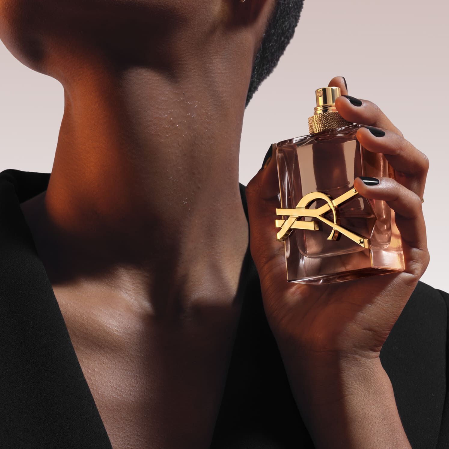 Gros plan: une main tient un flacon de parfum Libre en verre rectangulaire avec un logo YSL dore et un bouchon. La personne porte un blazer noir. Arriere-plan de lumiere orange et blanche.​