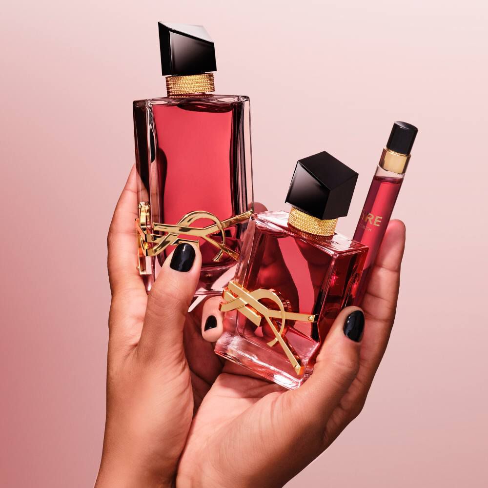 Mains aux ongles noirs tenant trois flacons de parfum YSL Libre de tailles differentes sur un fond rose.