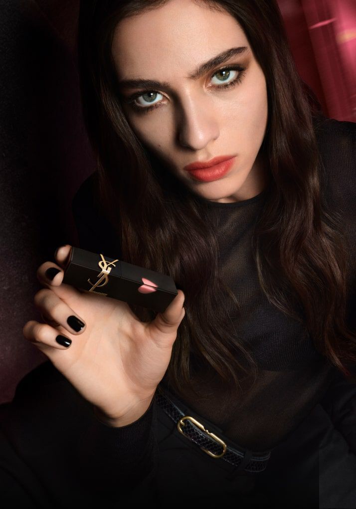 A person with long, dark hair and striking green eyes is holding a lipstick, looking directly at the camera with a bold expression. The background features warm, blurred hues, enhancing the dramatic feel of the image.