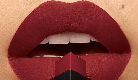Close-up of a pair of lips coated in deep red lipstick, with a red and black lipstick tube positioned just below.