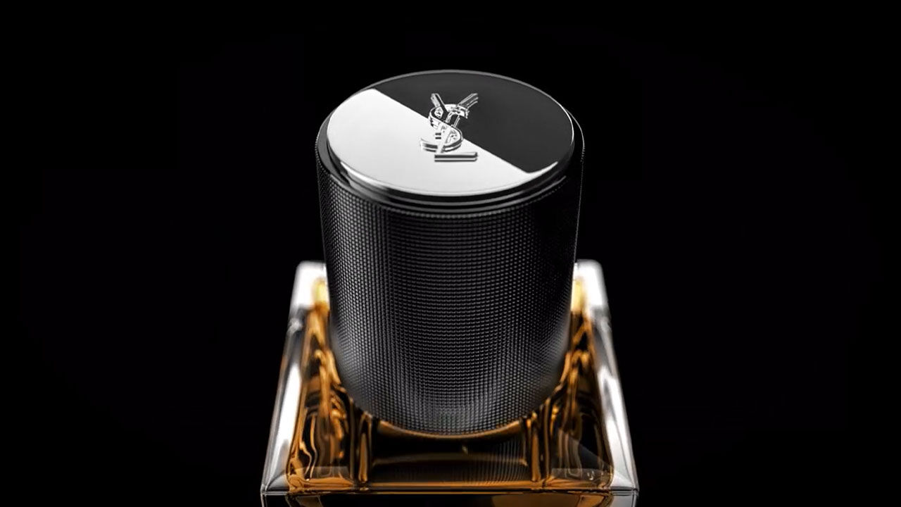 A sleek, black cylindrical perfume bottle with a shiny metallic cap, resting on a clear square base with amber liquid inside. The design features a subtle texture and a logo on top, set against a dark background.