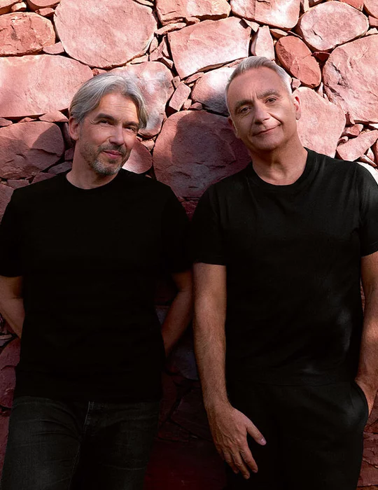 Deux hommes aux cheveux gris, tous deux vetus de t-shirts noirs, se tiennent adosses a un mur de pierre brun-rouge. L'homme de gauche est barbu et regarde vers le spectateur avec un leger sourire. L'homme de droite a les cheveux plus courts et regarde egalement vers le spectateur, une main dans la poche.