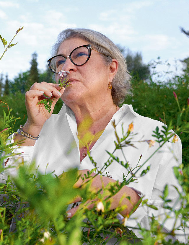 Une femme aux cheveux gris et aux lunettes noires est representee dans un jardin verdoyant, humant une petite fleur rose qu'elle tient pres de son nez. Elle porte une chemise blanche et des bijoux, notamment des boucles d'oreilles, un collier et des bracelets. Le ciel est d'un bleu eclatant, avec quelques nuages visibles a l'arriere-plan, ce qui suggere une journee claire. Le premier et le second plan sont occupes par diverses plantes vertes et quelques petites fleurs de couleur claire.