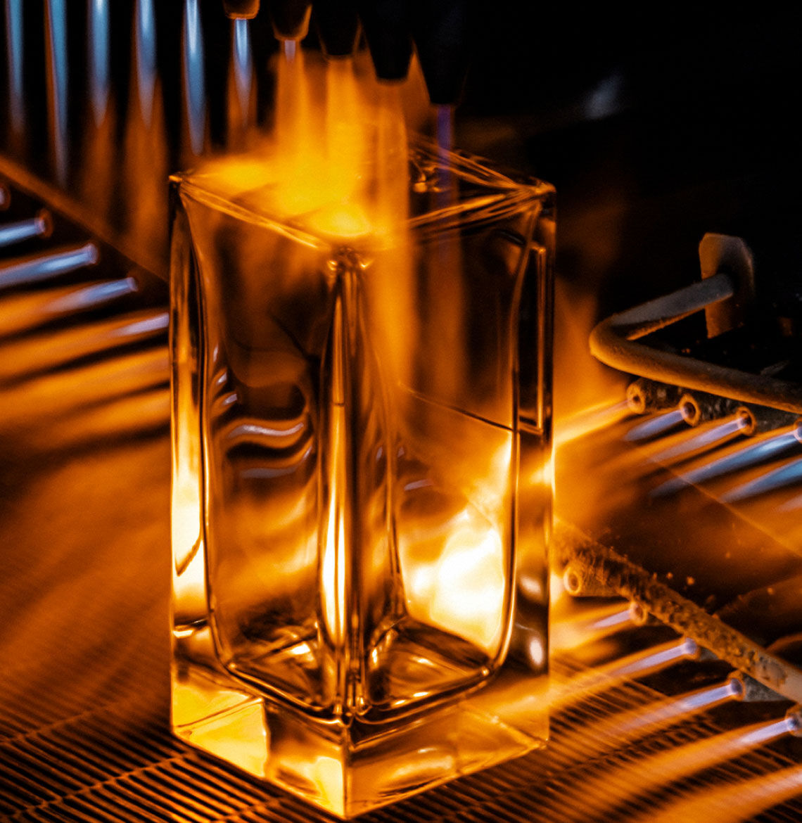 Une bouteille de parfum rectangulaire transparente est montree en train d'etre chauffee par des flammes provenant de plusieurs bruleurs, probablement dans le cadre d'un processus de traitement ou de formage du verre, creant une lueur chaude et ardente.