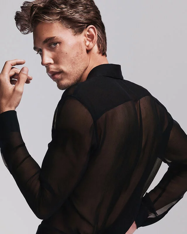 Un homme aux cheveux chatains clairs et aux yeux bleus, vetu d'une chemise noire transparente, regardant par-dessus son epaule.