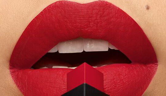 Close-up of a pair of perfectly shaped red lips, with a bold matte lipstick shade, holding a square lipstick tube against them.