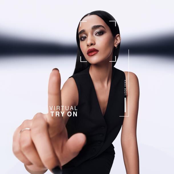 Une femme pointe en avant, interagissant avec un ecran d'essai virtuel. Un graphique affiche "VIRTUAL TRY ON" et "LOOK XXXX" autour de son visage. Elle porte un gilet noir.​