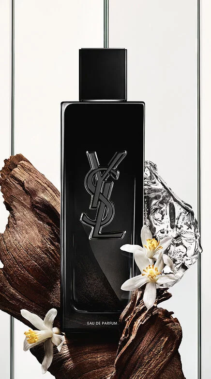 Une bouteille noire d'Eau de Parfum Yves Saint Laurent avec le logo YSL, entouree de morceaux de bois sombre, de fleurs blanches delicates et d'un objet reflechissant argente froisse.