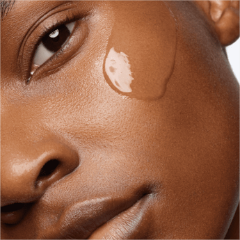 A close-up of a person's face with a smooth complexion, featuring a drop of foundation or skincare product applied to the cheek.