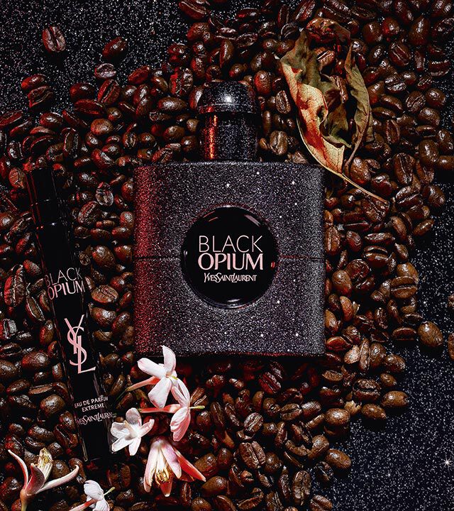 A dark, elegant perfume bottle labeled "Black Opium" by Yves Saint Laurent is surrounded by coffee beans and white flowers, creating a sophisticated and aromatic setting.