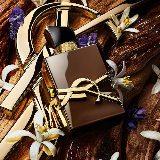 Flacon de parfum YSL Libre entoure de details dores, de fleurs d'oranger blanches et de fleurs mauves sur fond de bois.