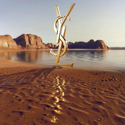 Un grand logo YSL (Yves Saint Laurent) dore en 3D pose sur un rivage desertique. Le monogramme dore poli reflete la lumiere du soleil et se trouve devant une etendue d'eau calme, avec des formations rocheuses rouges spectaculaires et des montagnes desertiques a l'arriere-plan sous un ciel degage.