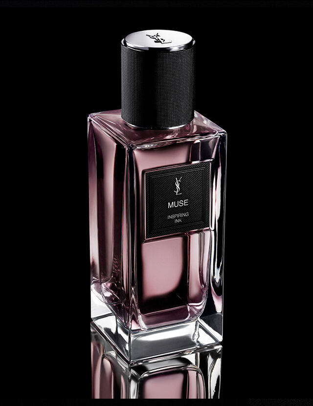 Une bouteille de parfum rectangulaire teintee de rose etiquetee "MUSE INSPIRING INK" de YSL, avec un bouchon noir texture, repose sur une surface reflechissante sur un fond sombre.