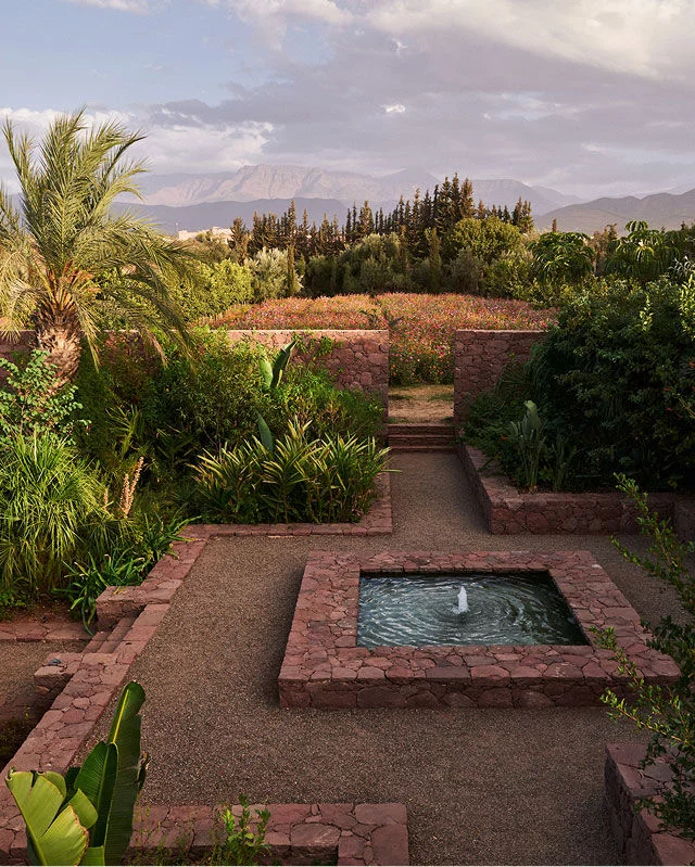 Un jardin verdoyant presente au premier plan une fontaine carree en pierre a l'eau bouillonnante, entouree d'allees en gravier et de murets en pierre. Des plantes et des arbustes verdoyants remplissent les plates-bandes du jardin, avec un champ de fleurs rouges et roses au milieu. A l'arriere-plan, des montagnes s'elevent sous un ciel doux et nuageux, et un grand palmier se dresse bien en evidence sur la gauche.