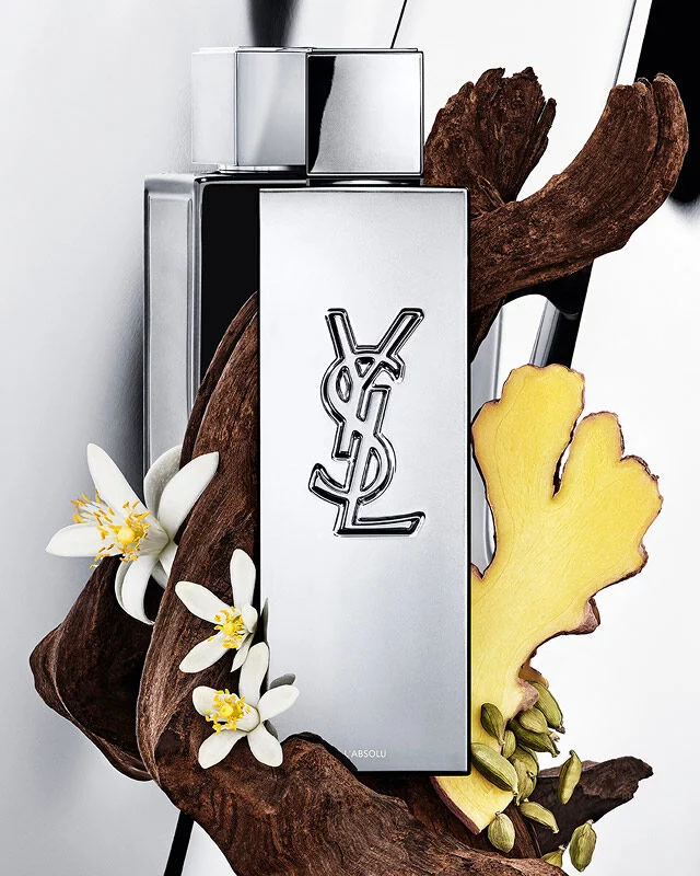 Flacon de parfum YSL Y L'ABSOLU entoure de bois fonce, de fleurs blanches de neroli, de gingembre et de gousses de cardamome.