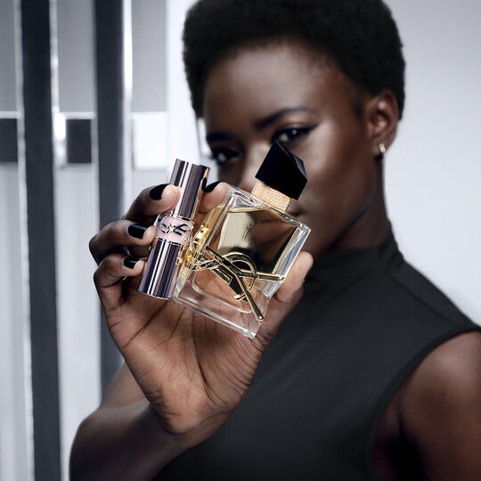 Une femme a la peau noire et aux cheveux courts tient un flacon de parfum Yves Saint Laurent et un rouge a levres assorti, tous deux ornes du logo YSL emblematique.