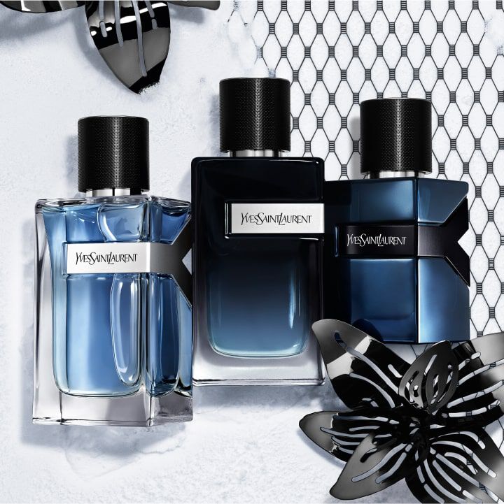 Three Yves Saint Laurent perfume bottles arranged against a textured white background, featuring sleek designs in shades of blue and black, accompanied by decorative black floral accents.