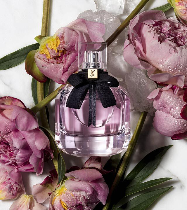 A clear perfume bottle with a black ribbon sits among pink peony flowers and ice on a marble surface.