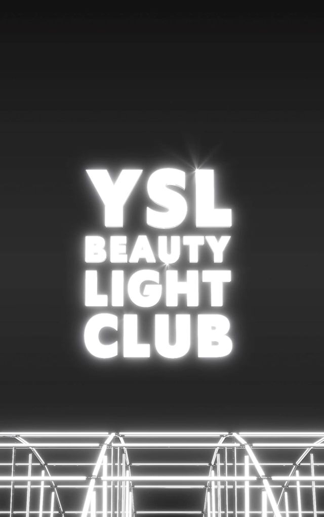 Un plan large et sombre revele le logo "YSL BEAUTY LIGHT CLUB" en lettres blanches lumineuses, flottant au centre d'un espace noir. En dessous du logo, une structure complexe de barres metalliques ou de neons blancs lumineux cree un effet de grille ou de scene, suggerant une installation lumineuse ou un environnement de club futuriste. L'ensemble degage une atmosphere moderne et chic.