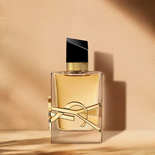 Un flacon de parfum en verre transparent avec un liquide dore, un bouchon noir et or, et un logo YSL dore bien visible, sur un fond chaud et neutre avec des ombres douces.