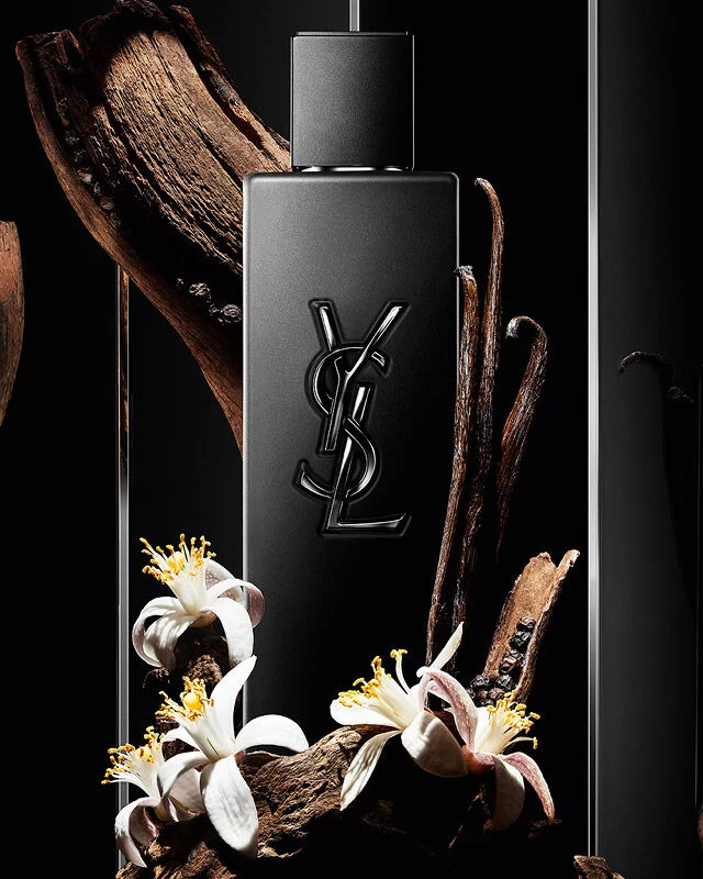 Flacon de parfum YSL noir entoure de bois, de gousses de vanille et de fleurs blanches.