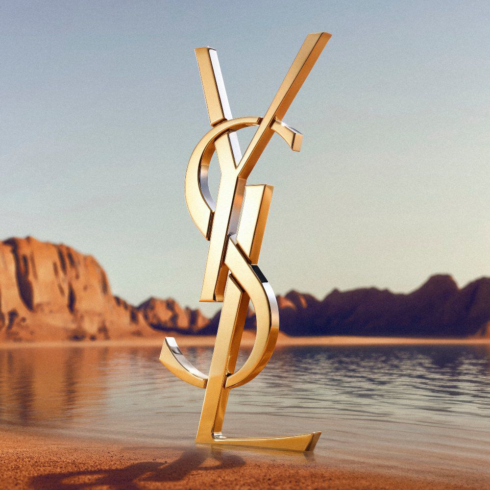Un grand logo YSL (Yves Saint Laurent) dore en 3D pose sur un rivage desertique. Le monogramme dore poli reflete la lumiere du soleil et se trouve devant une etendue d'eau calme, avec des formations rocheuses rouges spectaculaires et des montagnes desertiques a l'arriere-plan sous un ciel degage.​