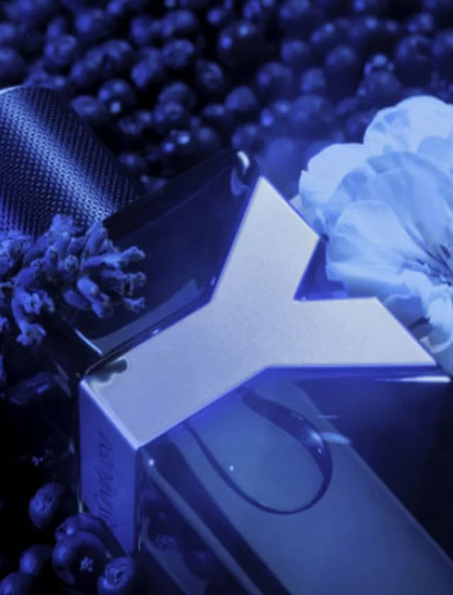 A close-up of a perfume bottle with a sleek design, surrounded by blueberries and flowers, all highlighted in soft blue lighting.