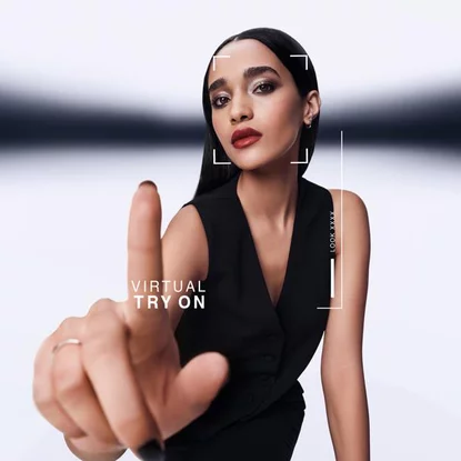Une femme pointe en avant, interagissant avec un ecran d'essai virtuel. Un graphique affiche "VIRTUAL TRY ON" et "LOOK XXXX" autour de son visage. Elle porte un gilet noir.