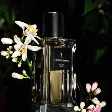 Une bouteille de parfum transparente etiquetee "SAHARIENNE SULTRY NEROLI" de YSL, placee sur un fond sombre avec de delicates fleurs de neroli blanches.