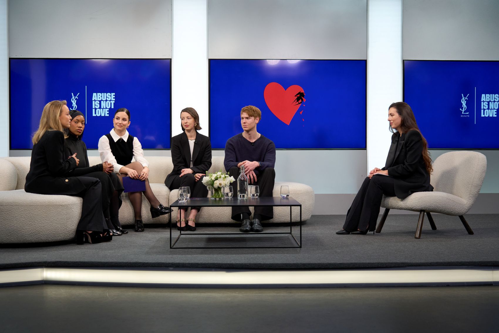 Table ronde avec six intervenants (quatre femmes, deux hommes) assis sur des canapes, devant de grands ecrans bleus affichant des messages, dans une salle moderne.