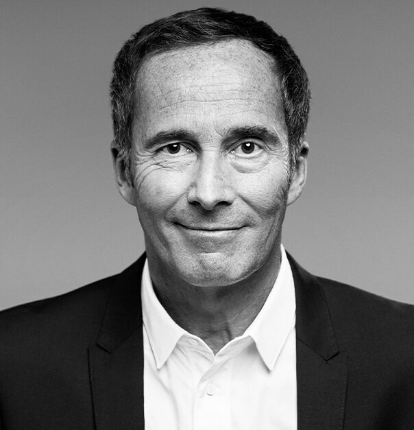 A smiling man with short, dark hair wearing a suit and a white shirt, against a neutral gray background.