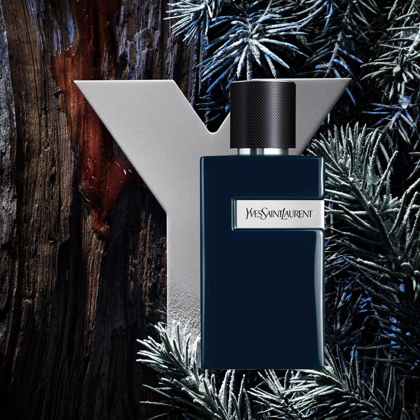 A stylish bottle of Yves Saint Laurent fragrance is set against a backdrop of textured wood and frosted pine foliage, with a prominent silver "Y" logo.
