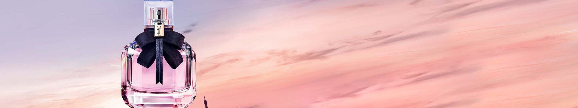 A perfume bottle with a black ribbon sits against a soft gradient sky at sunset, overlooking a cityscape.