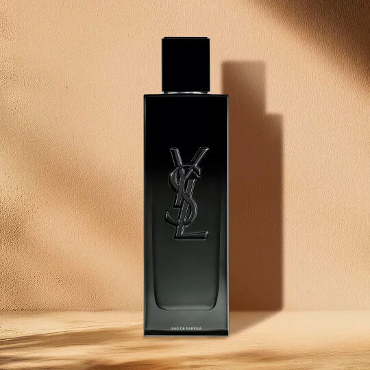 Un flacon de parfum noir, rectangulaire et elegant, avec un logo YSL noir en relief sur le devant. Les mots "EAU DE PARFUM" sont visibles pres du bas. Le flacon est muni d'un bouchon noir et se trouve sur une surface en bois clair, sur un fond beige uni et chaud, avec des ombres portees sur la droite.