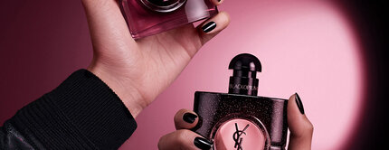 Deux mains tenant des flacons de parfum Yves Saint Laurent Black Opium sur un fond rose fonce et noir.