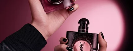 Deux mains tenant des flacons de parfum Yves Saint Laurent Black Opium sur un fond rose fonce et noir.