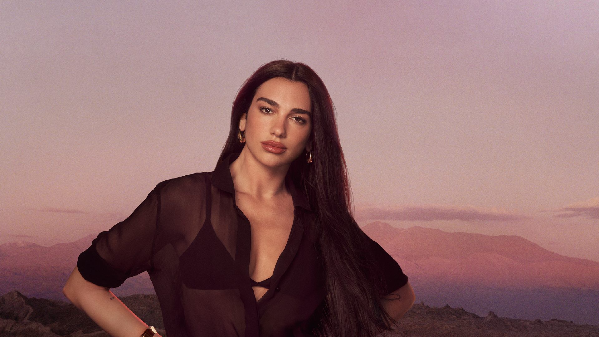 Dua Lipa posant en chemise noire transparente devant un paysage de montagnes au coucher du soleil.