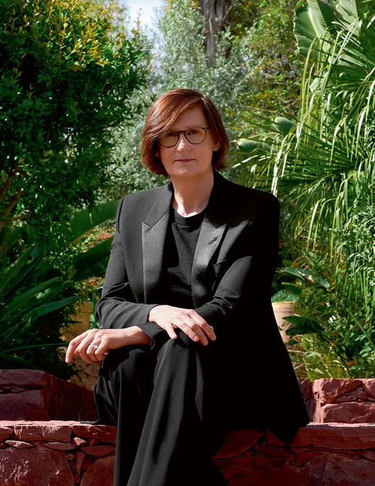 Une femme aux cheveux roux et aux lunettes est assise sur un mur de pierre, vetue d'un tailleur noir. Elle regarde le spectateur, avec un jardin verdoyant en arriere-plan.