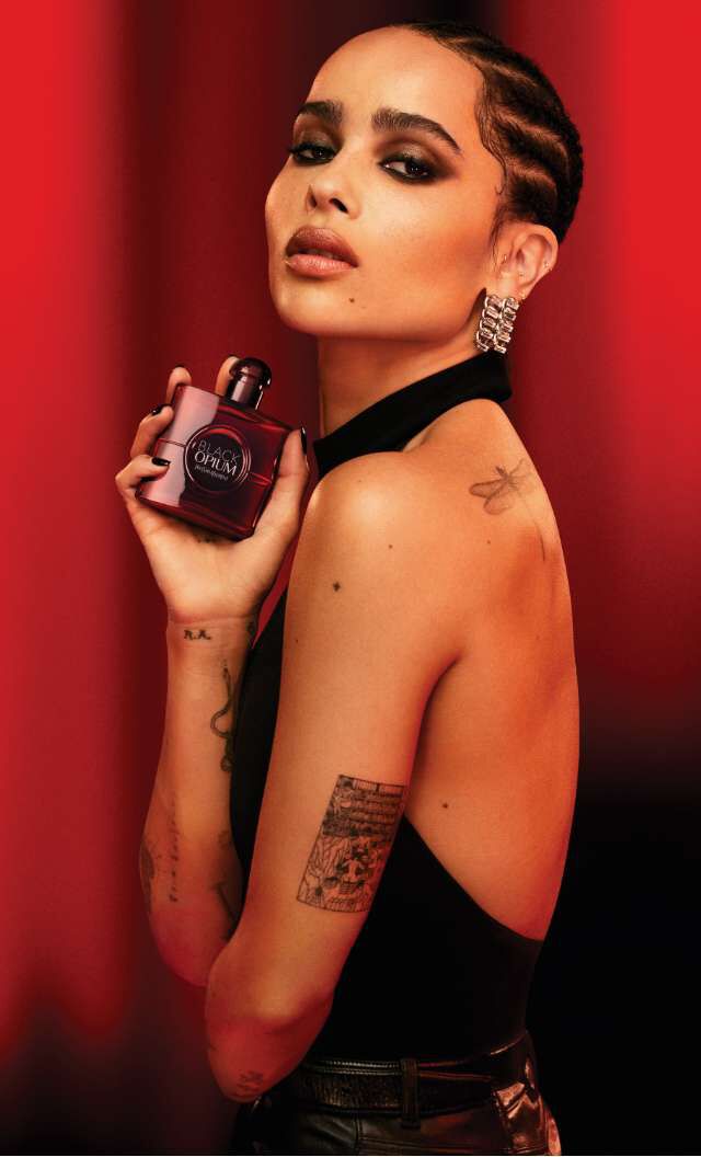 Une femme avec une robe sans manches, tenant un flacon de parfum, se tourne vers la camera sur un fond rouge, degageant une ambiance elegante et captivante.