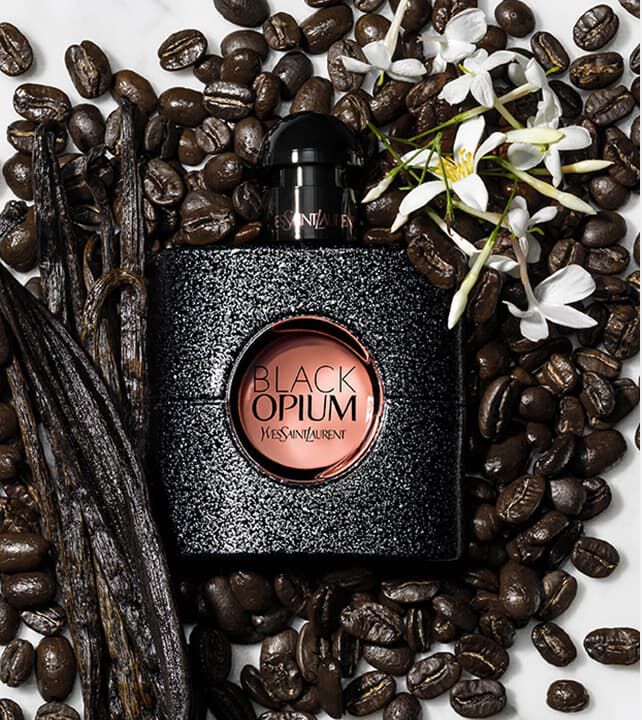 A bottle of Yves Saint Laurent Black Opium perfume surrounded by coffee beans and vanilla pods, with a white flower accenting the scene.