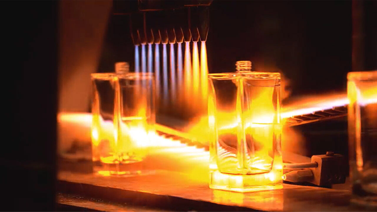 Gros plan d'une bouteille de parfum rectangulaire transparente chauffee intensement par des flammes provenant de plusieurs buses, soulignant le processus industriel avec une lueur orange.