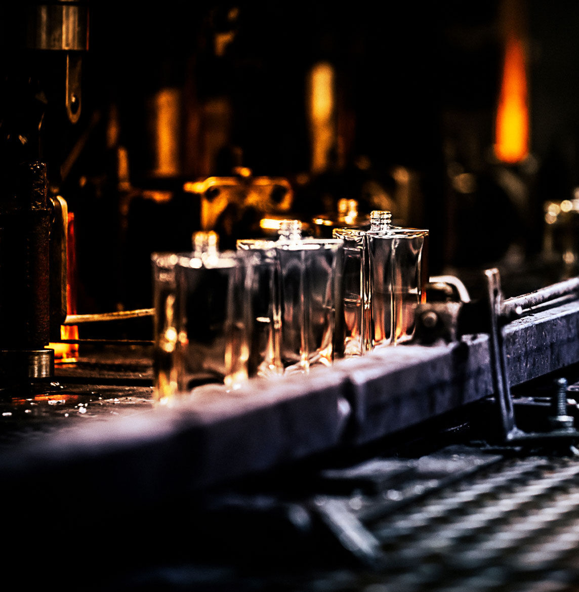 Plusieurs bouteilles de parfum transparentes, brillamment ambrees par l'arriere, sont alignees sur un tapis roulant ou une ligne de production dans une usine faiblement eclairee.