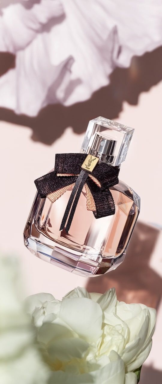 A stylish perfume bottle with a black ribbon and gold tag is suspended above soft white flowers, set against a pink background with subtle floral patterns.