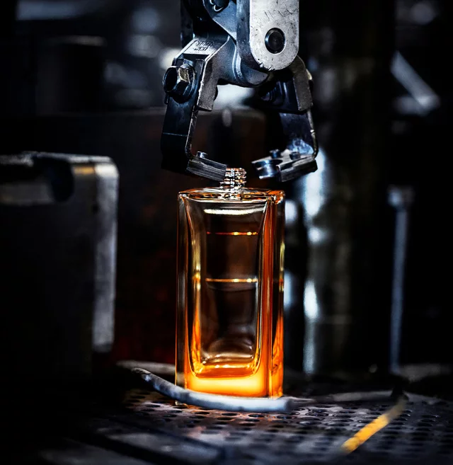 Un bras robotise abaisse un capuchon texture sur une bouteille de parfum ambree et lumineuse dans un cadre de fabrication, soulignant la precision de la production.