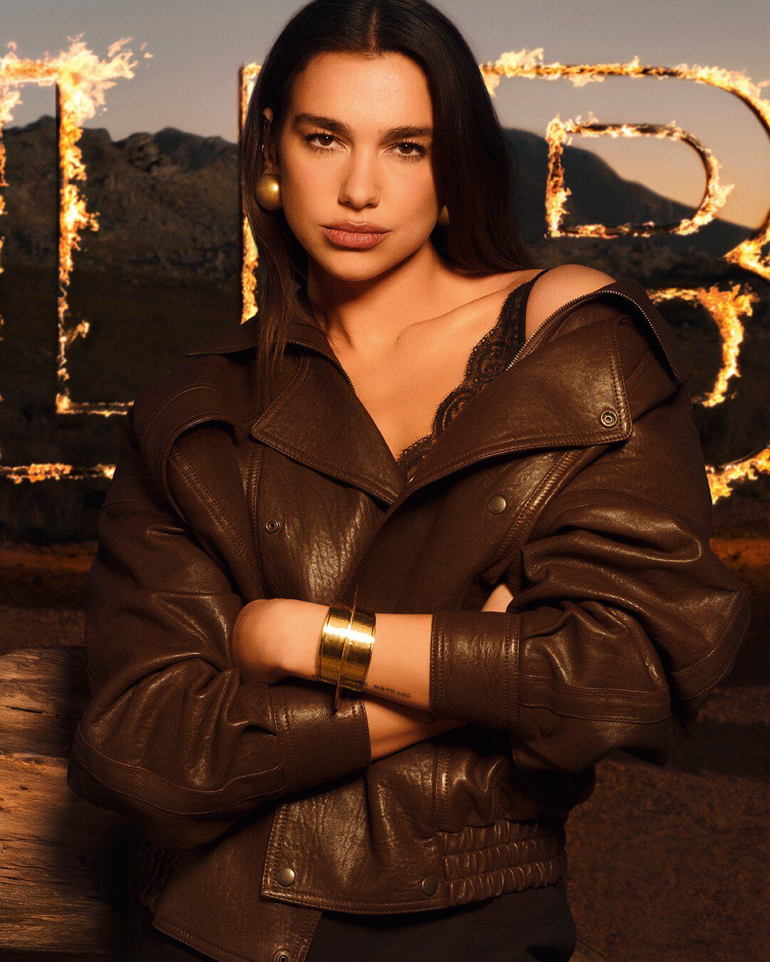 Dua Lipa dans une veste en cuir marron, devant de grandes lettres LIBRE enflammees, au coeur d’un paysage desertique brut.