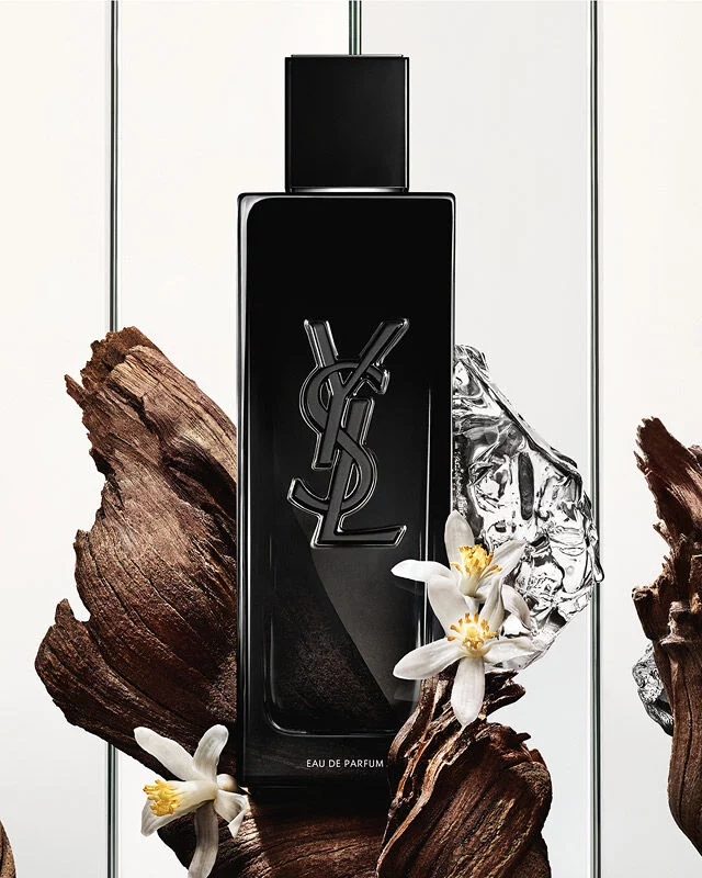 Une bouteille noire d'Eau de Parfum Yves Saint Laurent avec le logo YSL, entouree de morceaux de bois sombre, de fleurs blanches delicates et d'un objet reflechissant argente froisse.
