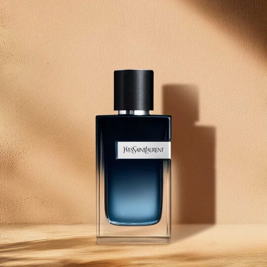 Flacon de parfum rectangulaire avec un liquide bleu fonce qui passe a un bleu plus clair vers le bas. Il est dote d'un bouchon noir texture, d'une bande argentee autour du col et d'une etiquette argentee sur le devant ou l'on peut lire "YVES SAINT LAURENT". Le flacon repose sur une surface en bois clair, avec des ombres douces derriere lui, sur un fond beige chaud.