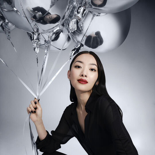 Une femme aux longs cheveux noirs, vetue d'une chemise noire, tient des ballons argentes et regarde directement l'objectif. Elle porte du rouge a levres rouge.