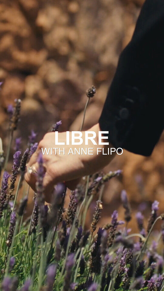 Une main, appartenant peut-etre a Anne Flipo, se tend vers un champ de fleurs violettes et lavande. La main porte une bague et est partiellement masquee par le texte superpose "LIBRE AVEC ANNE FLIPO" en blanc. A l'arriere-plan, on apercoit une paroi rocheuse floue.