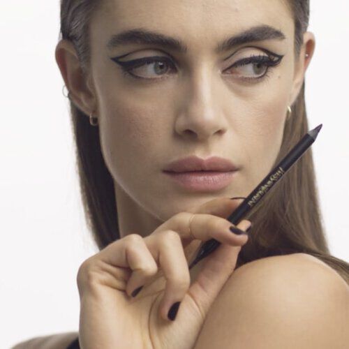 A model poses with a makeup pencil, showcasing bold winged eyeliner and a confident expression against a simple background.